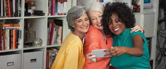 Three senior women laughing as they take a selfie together in front of a bookshelf. 